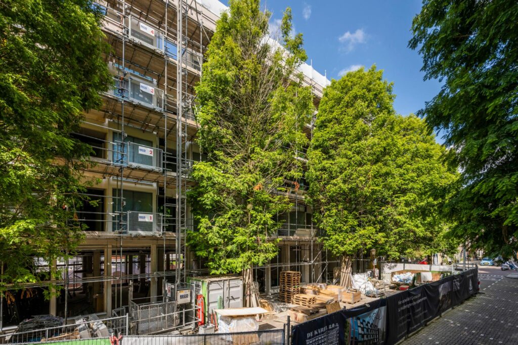 Een appartementencomplex aan de Karel du Jardinstraat is in renovatie, omringd door steigers en grote bomen.